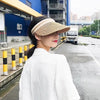 A woman wearing a white coat and a woven straw sun hat is standing on an asphalt road next to a blue building.