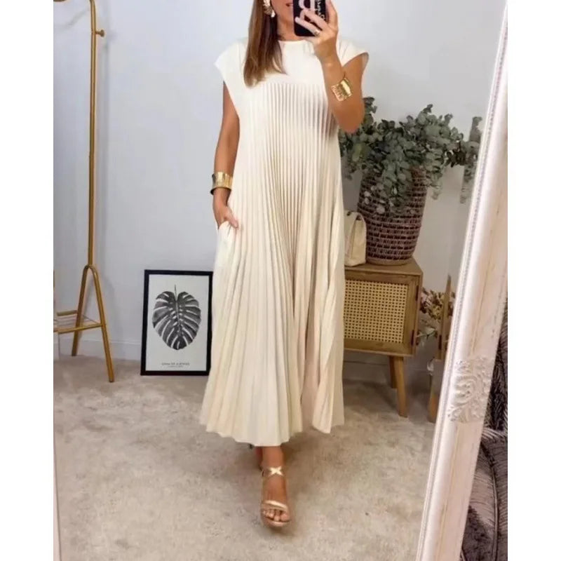 A woman wearing a cream pleated midi dress with a round neck standing in a bright room.