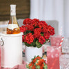 A bouquet of lifelike red artificial geraniums in a white pot next to a bottle of orange juice and pink drinks.