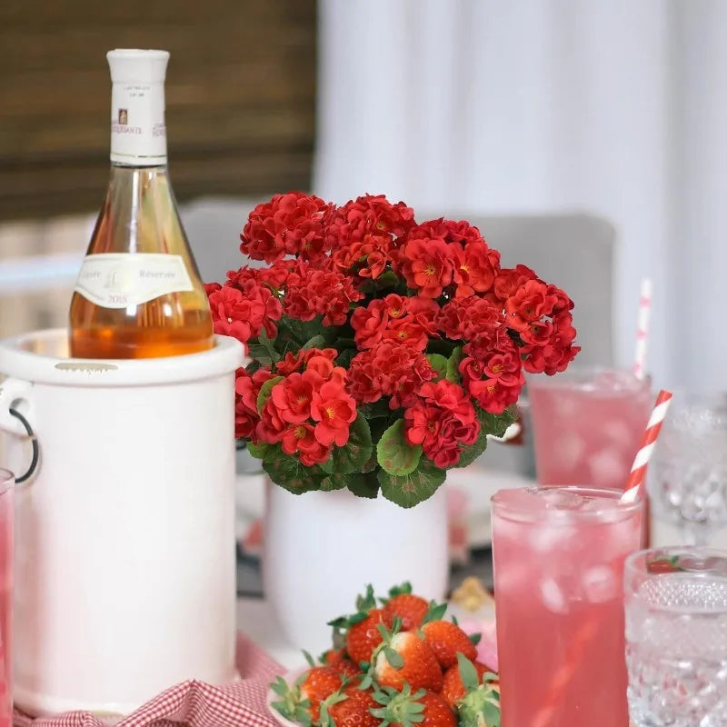 A bouquet of lifelike red artificial geraniums in a white pot next to a bottle of orange juice and pink drinks.