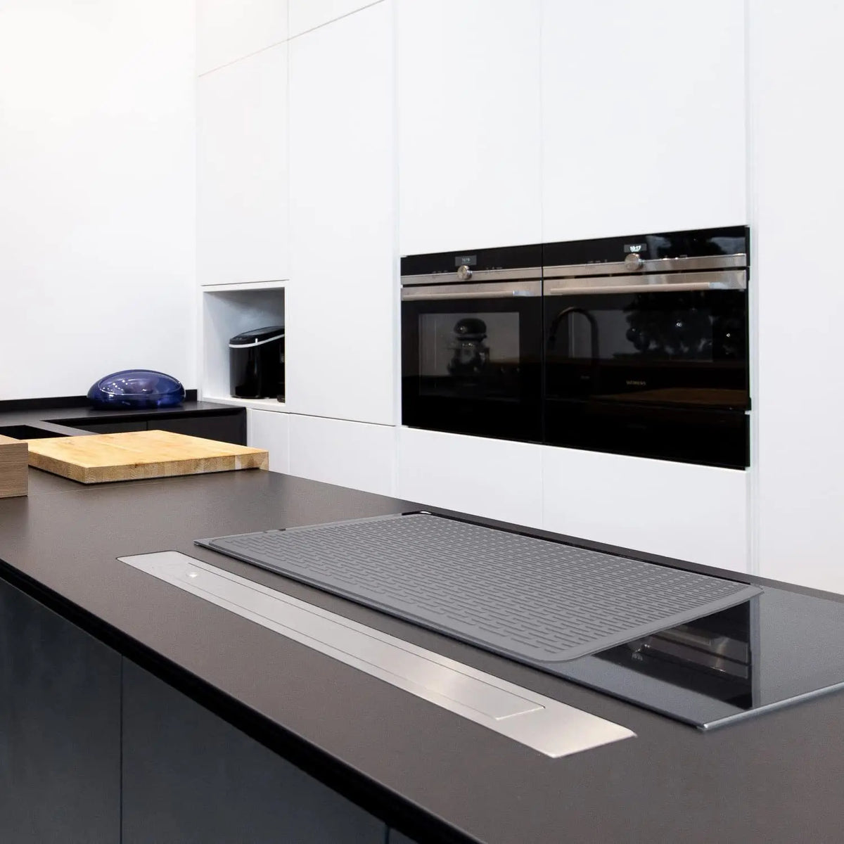 A black silicone stove protector mat is placed on a dark countertop in front of modern kitchen cabinetry.