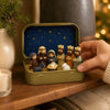 A hand opens a small gold-colored box revealing a miniature nativity scene with figures and animals against a dark blue starr