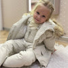 A young girl wearing a beige baby winter down jacket and pants set with a faux fur hood.