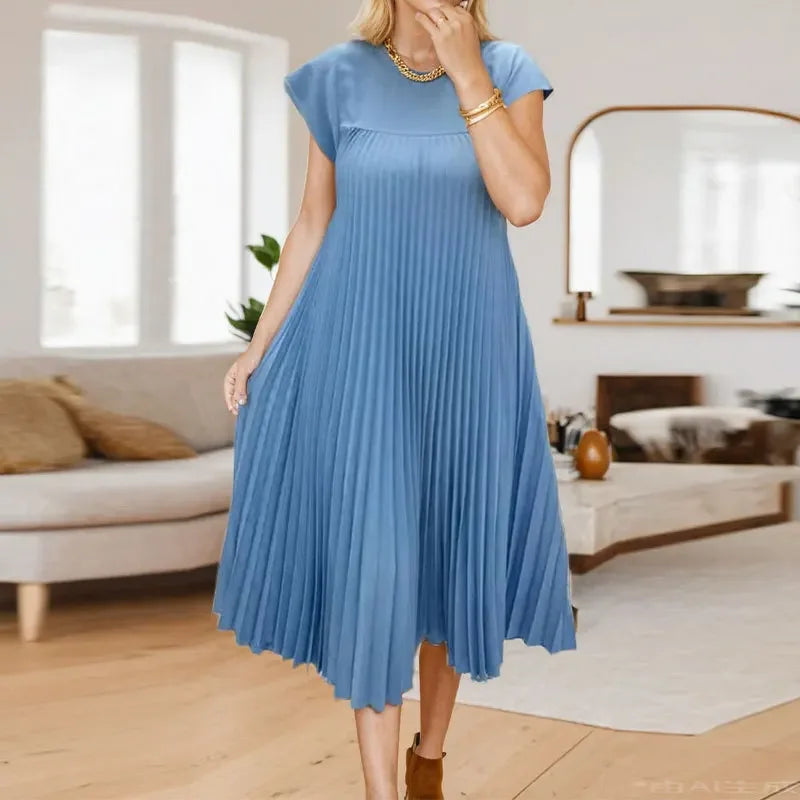 A woman wearing a dusty blue pleated midi dress with a round neckline standing indoors.