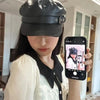 A woman wearing a black PU leather beret hat and a white top is holding a smartphone displaying a picture of a person.