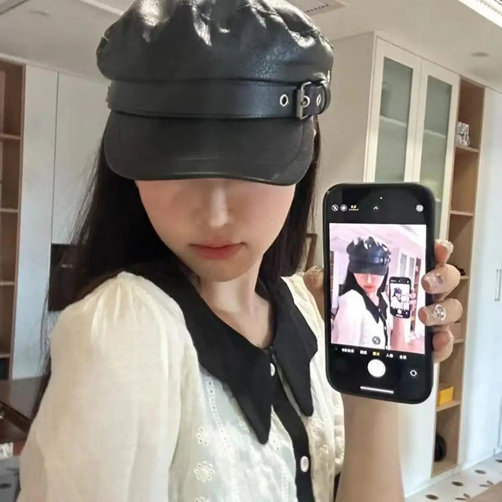 A woman wearing a black PU leather beret hat and a white top is holding a smartphone displaying a picture of a person.