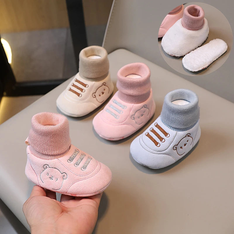 A hand holds a pair of pink knit baby booties with bear designs, shown next to other colored booties.