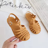 A pair of tan Roman gladiator jelly sandals with woven straps and a non-slip sole being held by a hand.