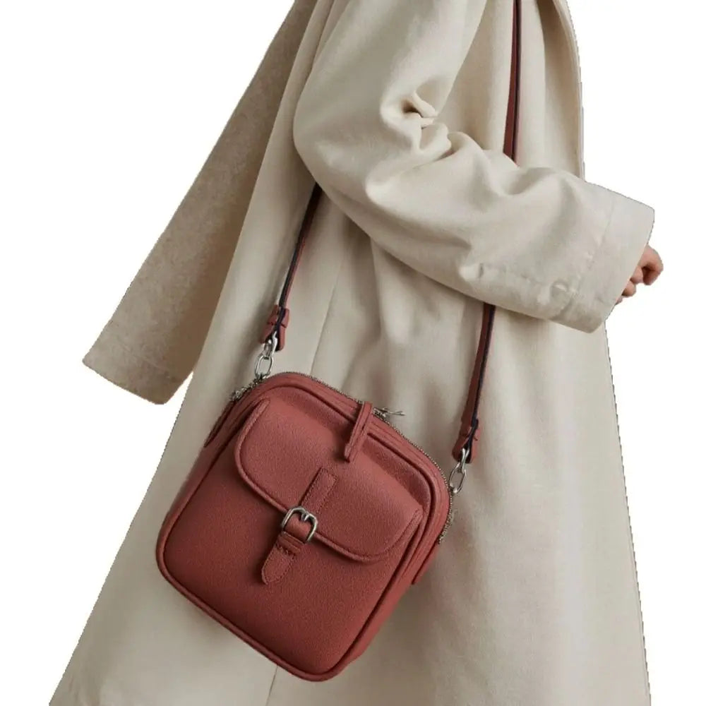A woman holds a dusty rose vegan leather messenger bag with a leather strap against a light beige coat.