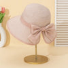A light pink straw sun hat with a large bow is displayed on a gold stand against a cream background.