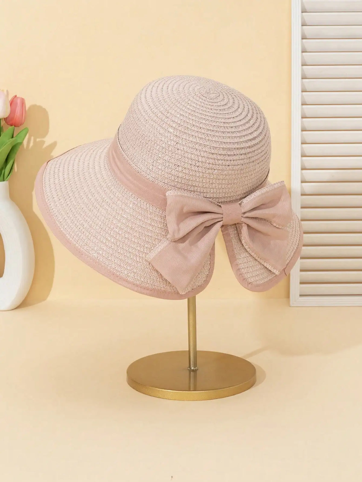 A light pink straw sun hat with a large bow is displayed on a gold stand against a cream background.