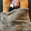 A close-up of a beige knit baby blanket with the name Baker Robert embroidered on it, resting next to a sleeping newborn.