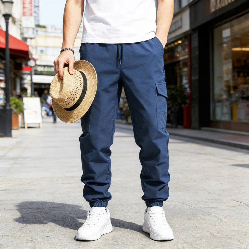 A man wearing dark blue casual cargo pants with a drawstring waist and multi-pockets standing on a sidewalk.