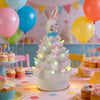 Figurine de lapin de Pâques en céramique blanche avec lumières LED, entourée de ballons colorés sur une table.