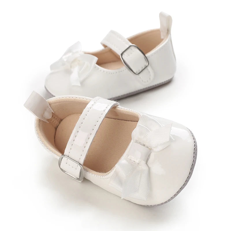 A pair of white soft non-slip baby first walker shoes with decorative bows.