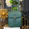 A dark green vegan leather messenger bag with gold hardware displayed on a wooden surface.