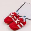 A pair of red baby first walker shoes with white laces and a sole, resting on a white background next to a card.