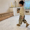 A young child wearing beige long-sleeved top and checkered brown harem pants is standing in a bright room.