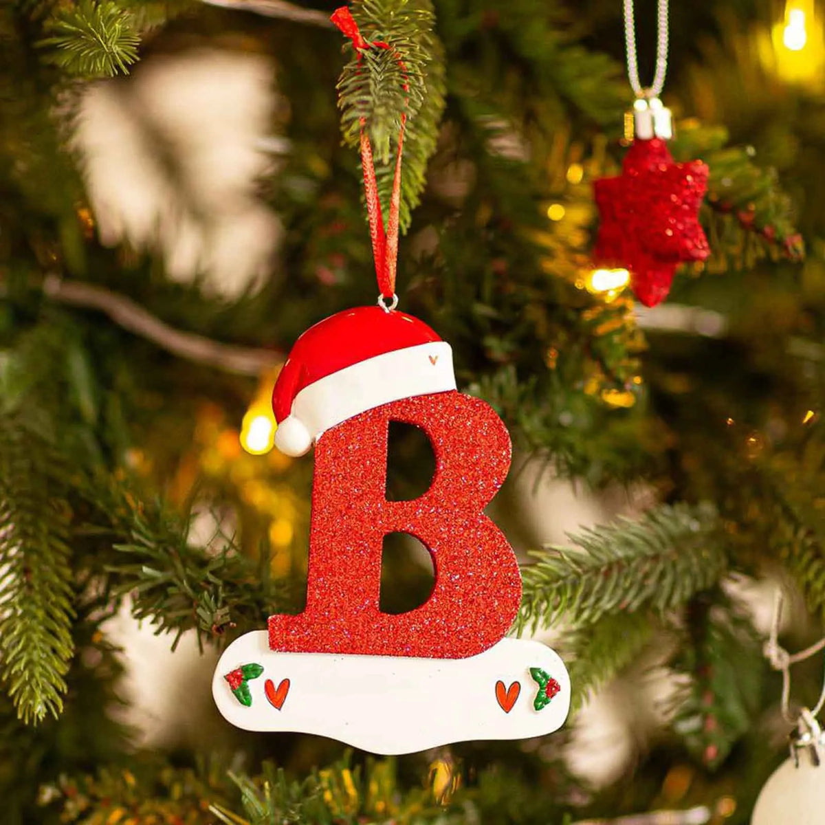 Rød glitter-akryl bokstav B på hvit base, hengende på et juletre.
