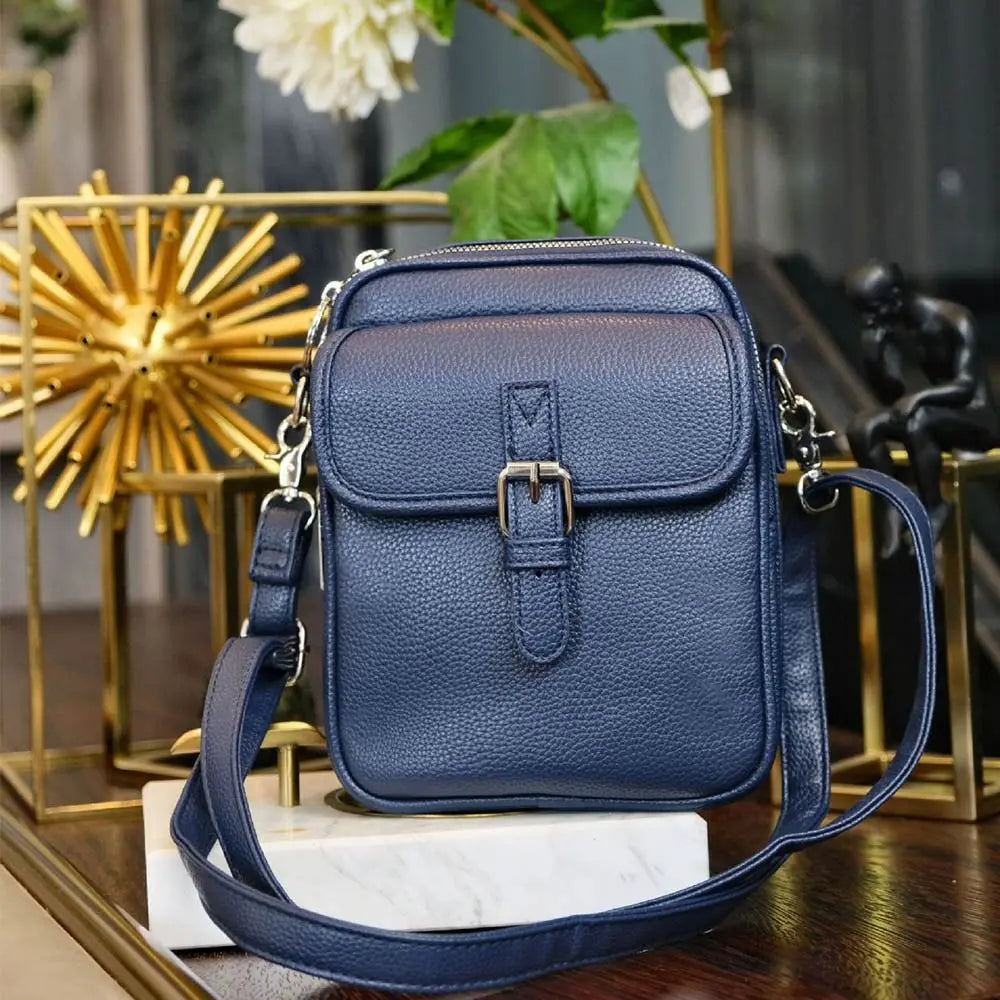 A navy blue vegan leather messenger bag with a gold buckle and adjustable strap resting on a wooden surface.