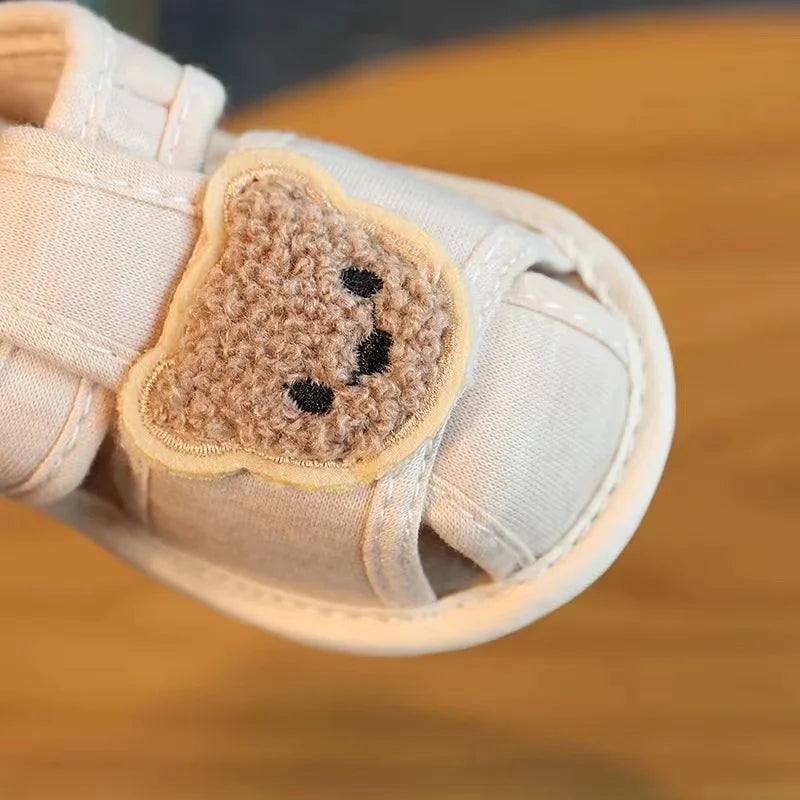 A pair of beige soft-sole sandals for a baby featuring a cute, fuzzy bear appliqué on the toe area.