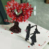 A 3D pop-up love tree card featuring a heart shape made of pink and red paper cutouts and a silhouette of a couple.