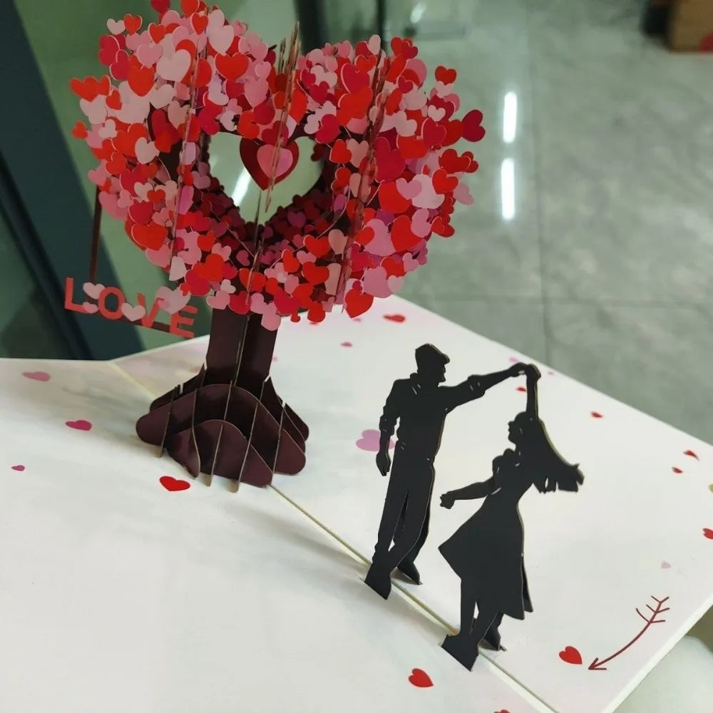A 3D pop-up love tree card featuring a heart shape made of pink and red paper cutouts and a silhouette of a couple.
