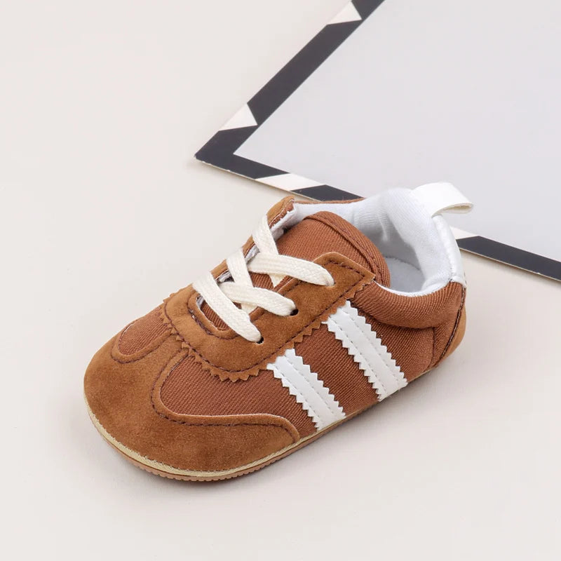 A pair of brown baby first walker shoes with white stripes and a canvas upper resting on a white background.
