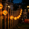 Halloween decor featuring string lights shaped like carved pumpkins and skulls decorating a wooden fence at dusk