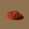 A close-up view of a burnt orange wool beret hat resting on a plain brown background.