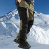 A person wearing olive green and black waterproof insulated fleece pants stands in a snowy mountain landscape.