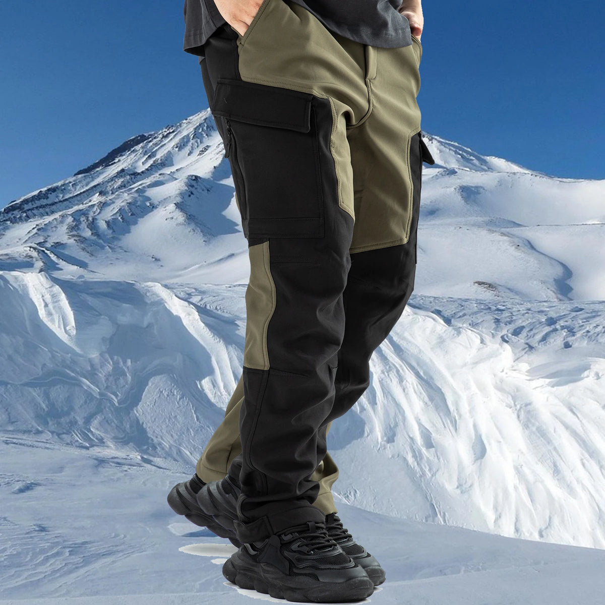 A person wearing olive green and black waterproof insulated fleece pants stands in a snowy mountain landscape.