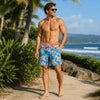 A muscular man wearing patriotic American flag print swim trunks stands outdoors near the ocean.