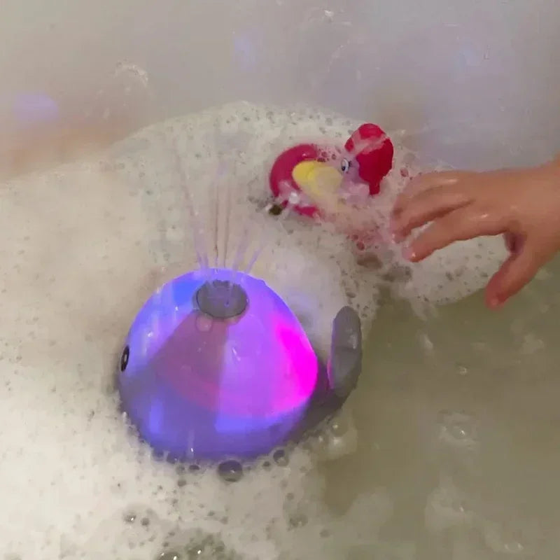 A hand is interacting with a purple musical whale bath toy that is spraying water and emitting light in a bathtub.