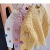 A close-up of a yellow knitted cardigan sweater with a textured stitch and pink pom-pom trim displayed on a hanger.