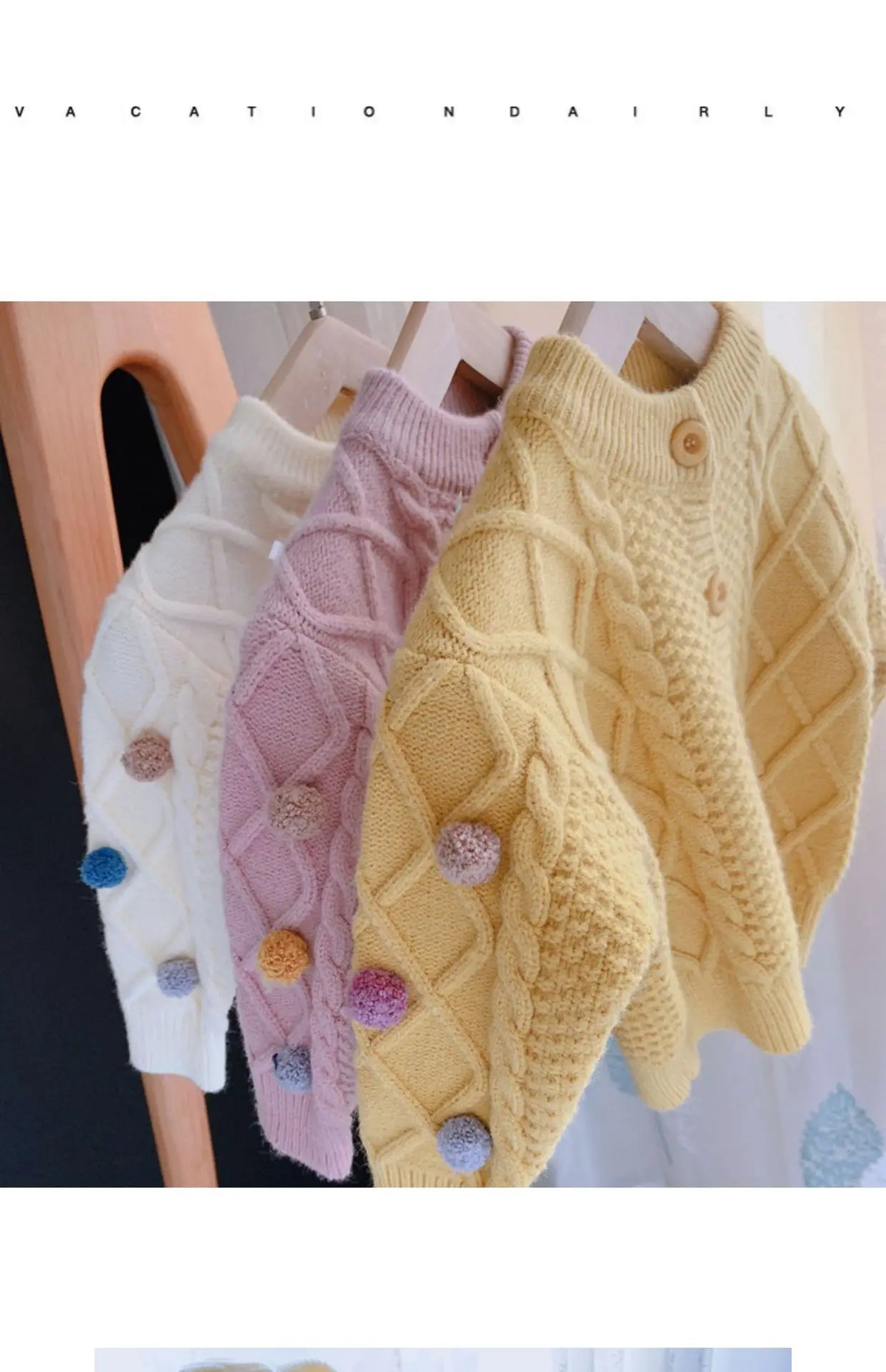 A close-up of a yellow knitted cardigan sweater with a textured stitch and pink pom-pom trim displayed on a hanger.