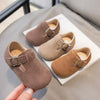 A hand holds a pair of brown suede kids' moccasin shoes with hook-and-loop closures on a white surface.