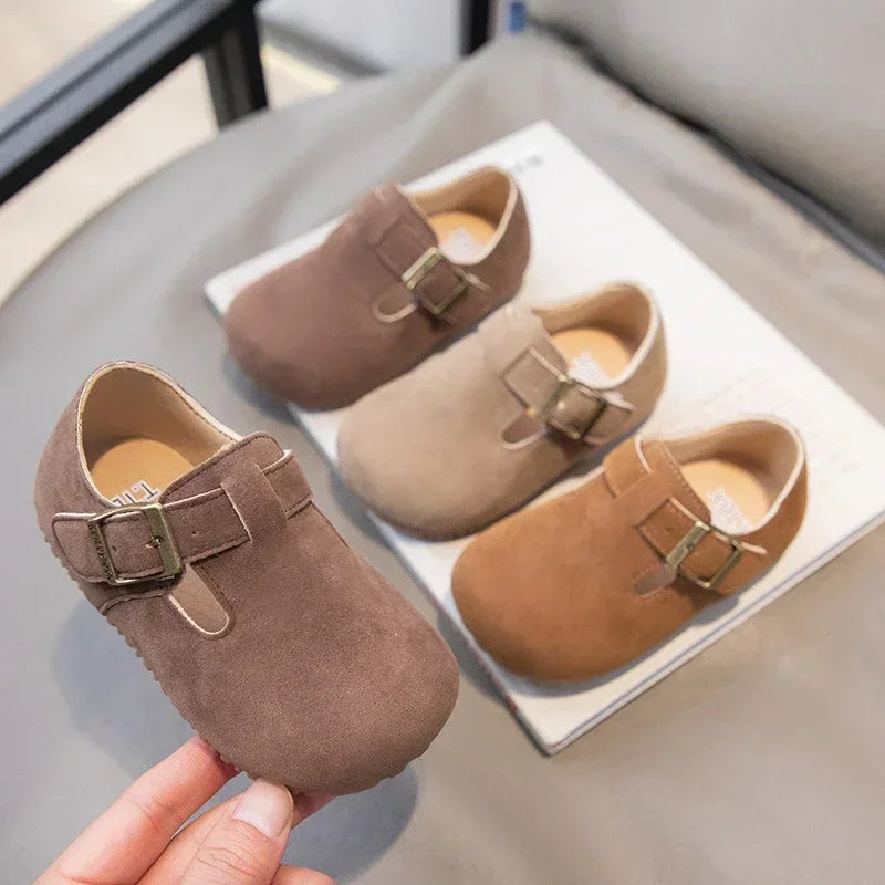 A hand holds a pair of brown suede kids' moccasin shoes with hook-and-loop closures on a white surface.