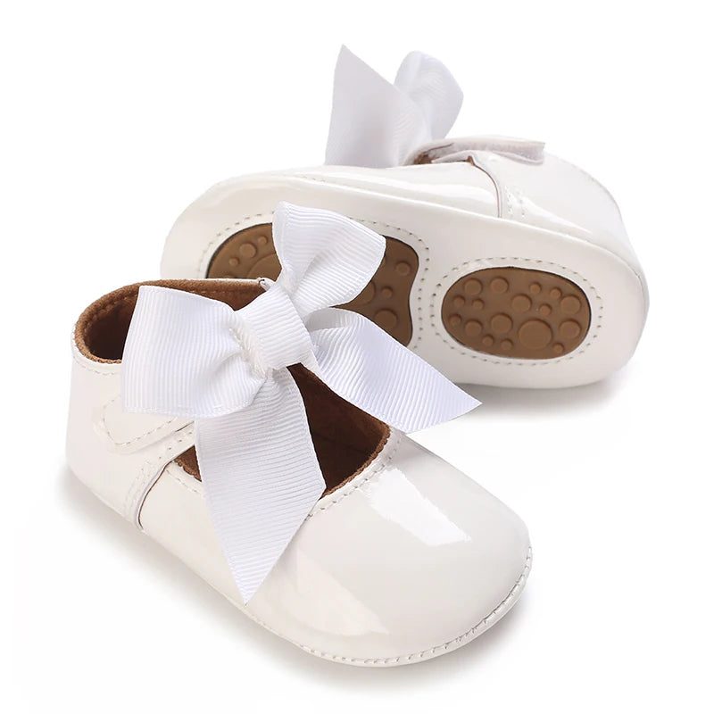A pair of white baby first walker shoes with white bows and brown non-slip soles.
