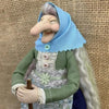 A handcrafted kitchen witch doll with a light blue hood and green dress is shown against a woven background.
