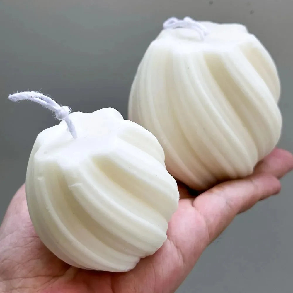 A hand holds two white pillar candles molded in a geometric wave pattern with white string attached.
