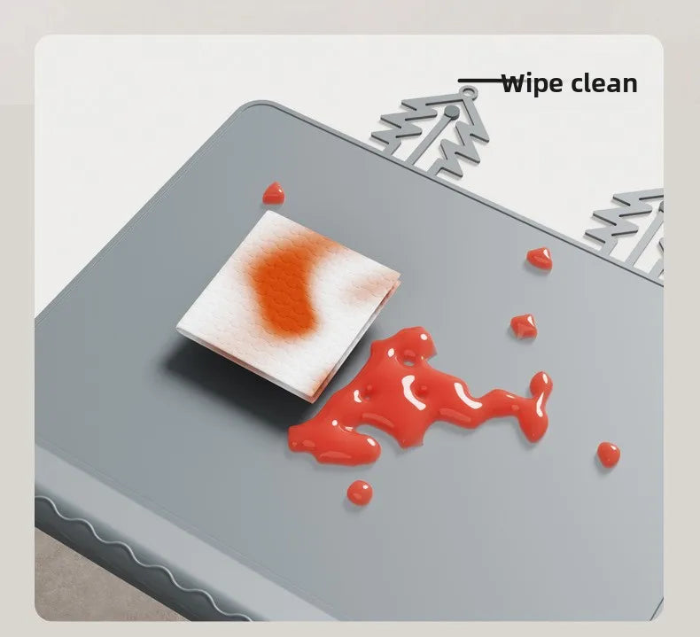 A silicone feeding placemat is shown with a reddish stain being wiped clean by a white cloth, demonstrating its cleaning abil