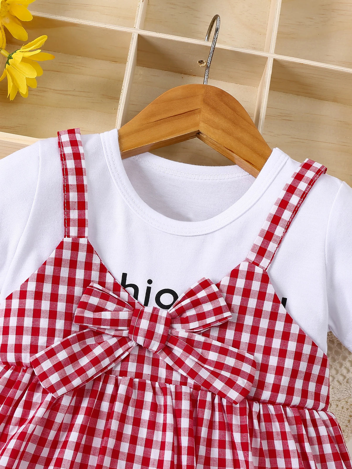 A red and white plaid girls' cotton knit dress with a bow accent displayed on a wooden hanger.