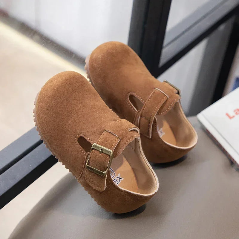 A pair of brown suede kids' moccasin shoes with an easy hook-and-loop closure resting on a dark surface.