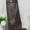 A pair of brown leather knee-high chunky heel boots with a metal buckle displayed on a stand.