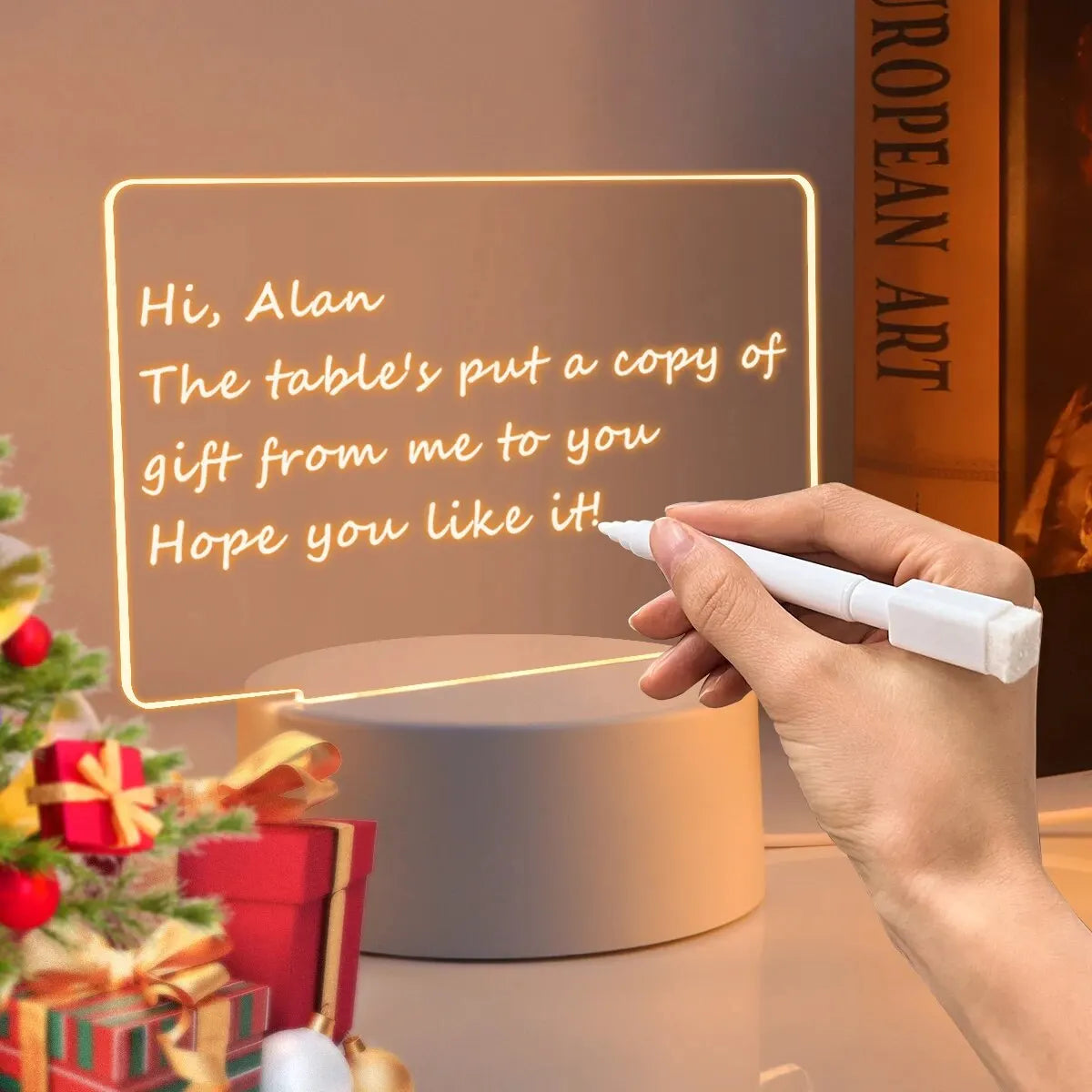 A hand writes a message on a glowing acrylic board with a pen in front of a desk lamp.