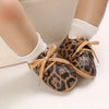 A pair of leopard print baby first walkers with soft rubber soles shown on a person's foot.