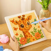 Hands are using a needle to embroider an orange giraffe pattern on a canvas within a wooden frame.