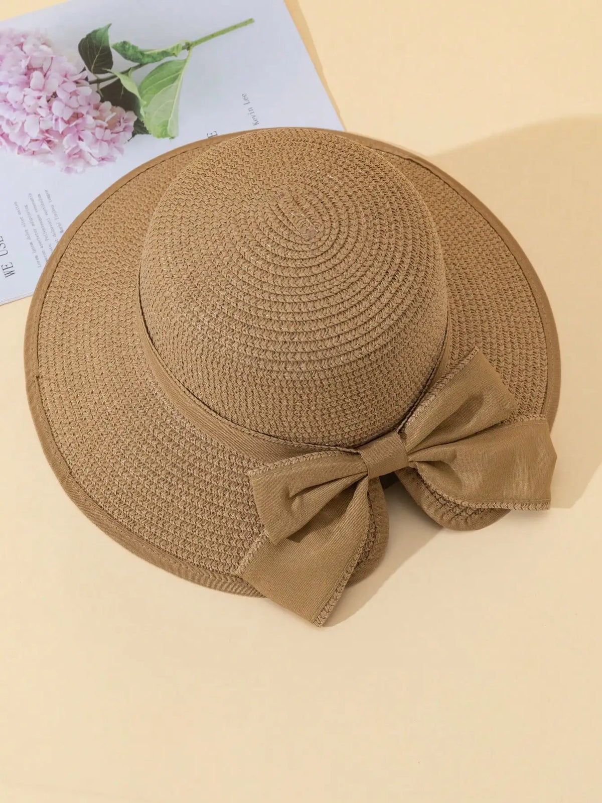 A beige straw sun hat with a wide brim and a matching bow, resting on a light background.