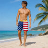 A muscular man wearing patriotic American flag print swim trunks stands on a beach with palm trees and blue water.